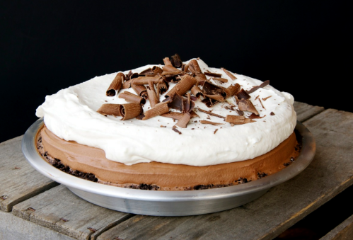 A chocolate cream pie topped with whipped cream and chocolate shavings, served in a silver pie dish on a wooden surface.