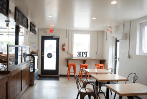A cozy café interior with wooden tables, orange stools, and a display case, featuring a sign that says "PIE."