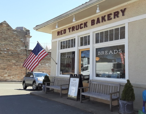 A quaint bakery with an American flag, benches outside, and a sign that says "We are OPEN!" on a sunny day.