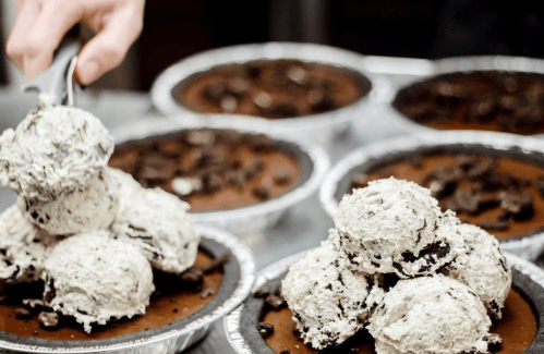 Scoops of cookies and cream ice cream on chocolate pies, with a hand serving from a nearby pie.