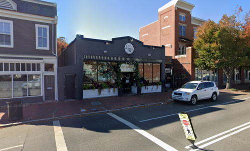 A street view of a storefront with large windows, surrounded by trees and a parked car in front.