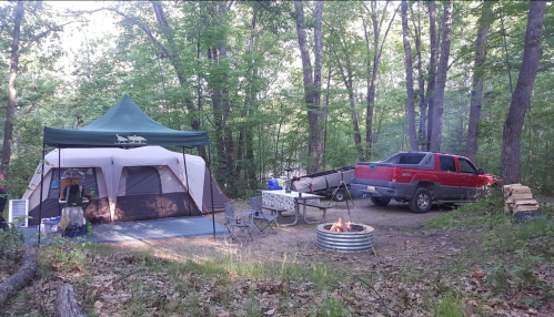 A campsite in the woods featuring a tent, picnic table, fire pit, and a red truck parked nearby.