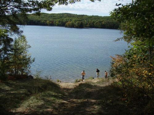 Three people stand by a calm lake surrounded by trees, with a clear sky and colorful foliage in the background.