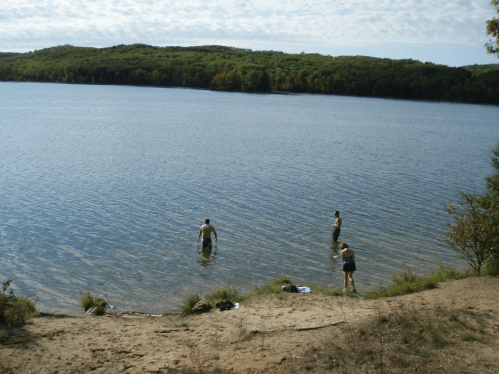 Two people wade in a calm lake, surrounded by trees and a sandy shore under a partly cloudy sky.