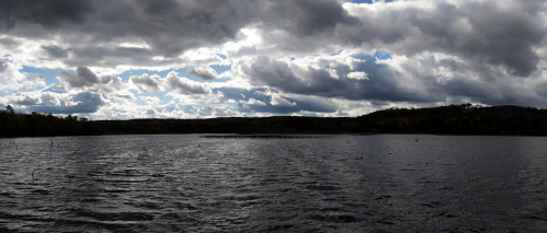 A serene lake under a dramatic sky filled with clouds, surrounded by trees and distant hills.