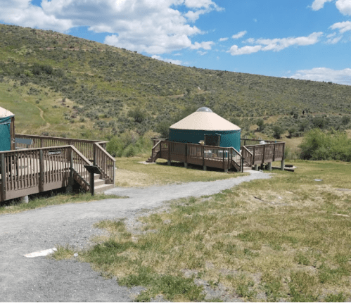 Two green yurts with wooden decks on a grassy path, surrounded by rolling hills and a blue sky with clouds.