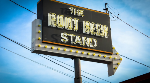 Sign for "The Root Beer Stand" with bright lights and an arrow, set against a blue sky.