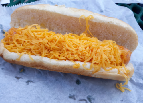 A hot dog bun filled with a crispy fried item and topped with a generous amount of shredded cheddar cheese.