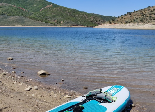 A paddleboard rests on a sandy shore by a calm lake, surrounded by green hills under a clear blue sky.