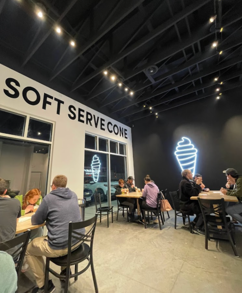 Interior of a modern ice cream shop with customers seated at tables, featuring a neon soft serve cone sign on the wall.