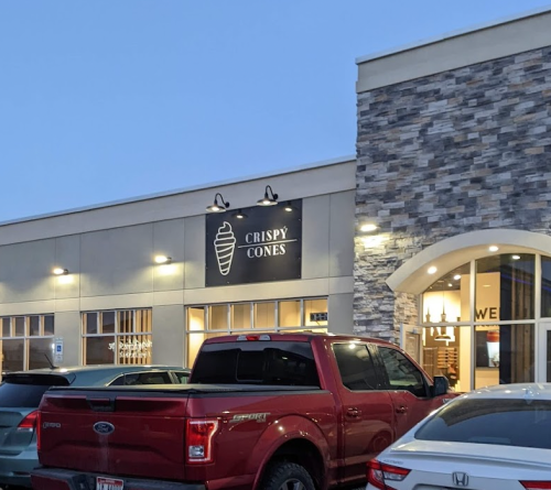 Exterior of "Crispy Cones" ice cream shop with a stone facade and parked cars in front during twilight.