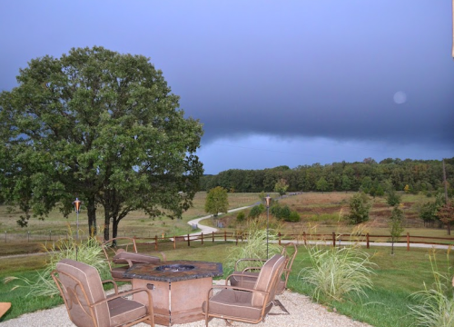 A stormy sky looms over a rural landscape with a fire pit and chairs in the foreground, surrounded by trees and a winding path.