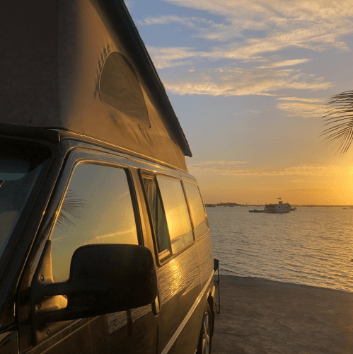 A van parked by the water at sunset, with palm trees and a distant boat on the horizon.