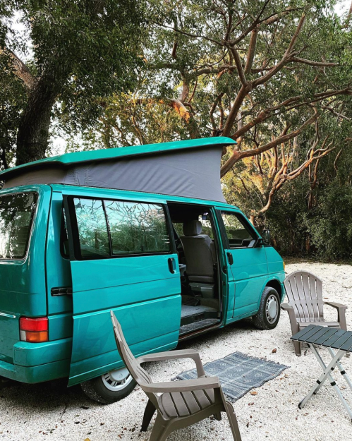 A teal camper van parked in a wooded area, with a pop-up roof and two chairs nearby on a gravel surface.