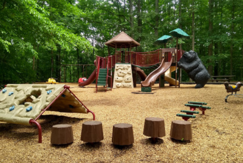 A playground with a climbing structure, slide, bear statue, and log seating, surrounded by trees and gravel ground.