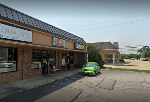 A storefront featuring a pie shop and a Subway, with a green car parked outside on a sunny day.