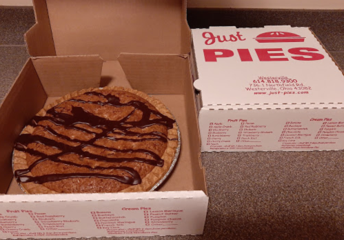 A pie with chocolate drizzle sits in an open box labeled "Just PIES," with a list of pie options on the side.