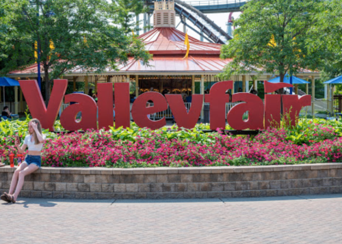 A young woman sits by a colorful "Valleyfair" sign surrounded by flowers and greenery at an amusement park.
