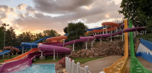 Colorful water slides twist down a hillside beside a swimming pool, with a sunset sky in the background.