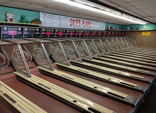 A row of skee-ball machines in an arcade, with scoreboards and colorful plush toys on display above.