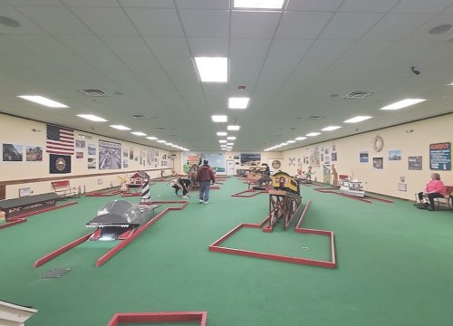 Indoor mini-golf course with green carpet, various obstacles, and players enjoying the game. Walls decorated with posters.