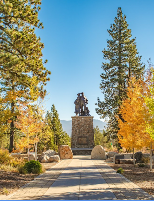 A statue on a stone pedestal surrounded by trees and colorful autumn foliage under a clear blue sky.