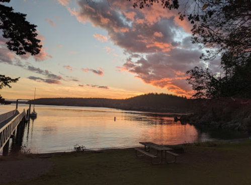 A serene sunset over a calm bay, with clouds reflecting pink and orange hues, and a picnic table in the foreground.