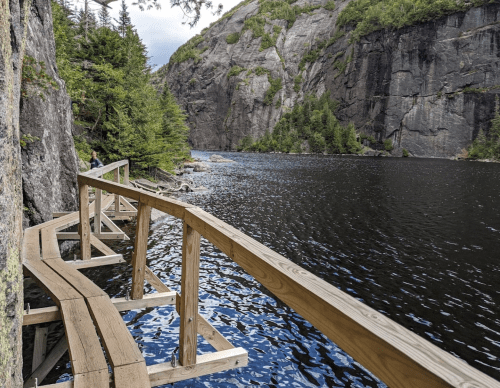 A wooden walkway curves along a calm lake, surrounded by steep rocky cliffs and lush greenery.