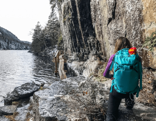 A person with a teal backpack sits on a rock by a lake, surrounded by trees and a rocky cliff.