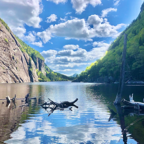 A serene lake surrounded by lush green hills and rocky cliffs, reflecting fluffy clouds in a bright blue sky.