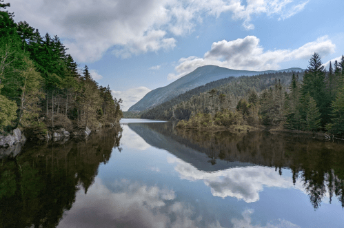 A serene lake surrounded by lush trees and mountains, reflecting the sky and clouds in calm waters.