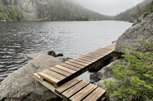 A wooden dock extends over a calm lake, surrounded by rocky shores and misty forested hills in the background.