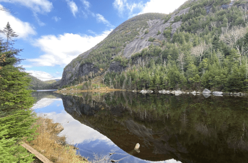 A serene lake reflects surrounding mountains and trees under a partly cloudy sky.