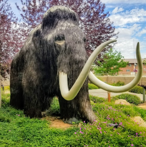 A large, lifelike woolly mammoth statue surrounded by greenery and purple flowers under a blue sky.
