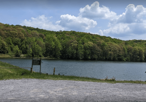 A serene lake surrounded by lush green trees under a partly cloudy sky, with a gravel area in the foreground.