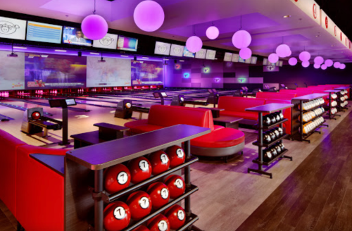A vibrant bowling alley with red seating, purple lighting, and multiple lanes, featuring red bowling balls on display.