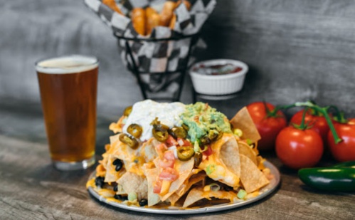 A plate of loaded nachos topped with cheese, jalapeños, guacamole, and sour cream, alongside a glass of beer and tomatoes.