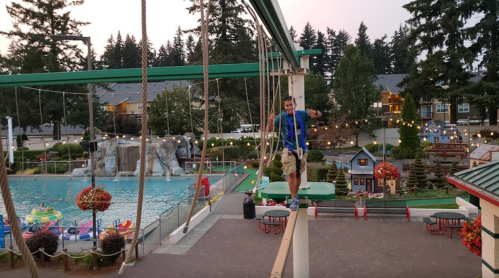A person balances on a rope bridge above a pool area with trees and colorful decorations in the background.
