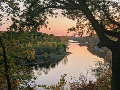 A serene river at sunset, surrounded by trees with autumn foliage, reflecting warm colors in the water.