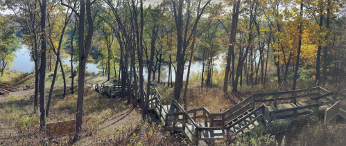 A winding wooden staircase descends through trees to a serene lake, surrounded by autumn foliage.