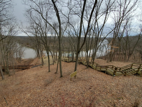 A winding wooden staircase descends through bare trees to a river, surrounded by fallen leaves and a cloudy sky.