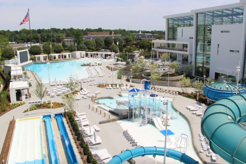 Aerial view of a modern pool area with slides, lounge chairs, and a large building in the background, under a clear sky.