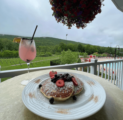 A plate of pancakes topped with strawberries and blueberries, alongside a pink drink, with a scenic view of green hills.