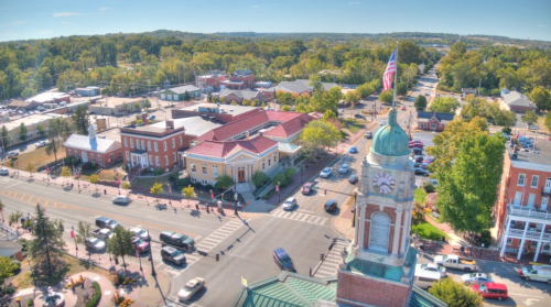 Aerial view of a town square with trees, buildings, and a clock tower, bustling with cars and pedestrians on a sunny day.