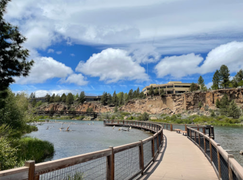 A winding wooden path along a river, surrounded by greenery and cliffs under a partly cloudy sky.