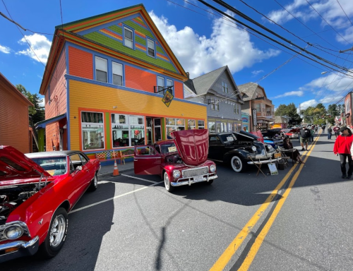 Colorful buildings line a street with classic cars on display during a car show under a blue sky with fluffy clouds.