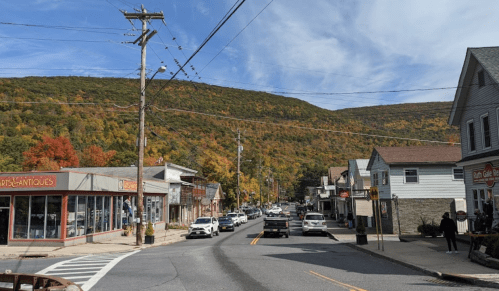 A quaint street lined with shops and cars, set against a backdrop of colorful autumn foliage on the mountains.
