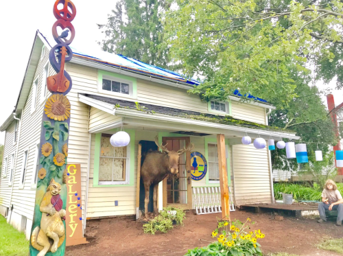 A quirky gallery with a wooden sign, featuring a moose at the entrance and a person sitting nearby.