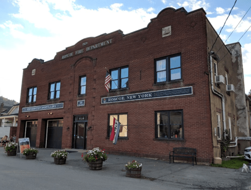 Historic brick building housing the Roscoe Fire Department in Roscoe, New York, with flower planters and an American flag.