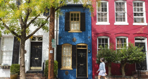 A person walks past colorful row houses: one white, one blue, and one red, with trees and greenery in the foreground.